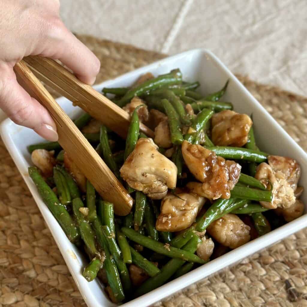 Grabbing with tongs a serving of green beans and chicken.