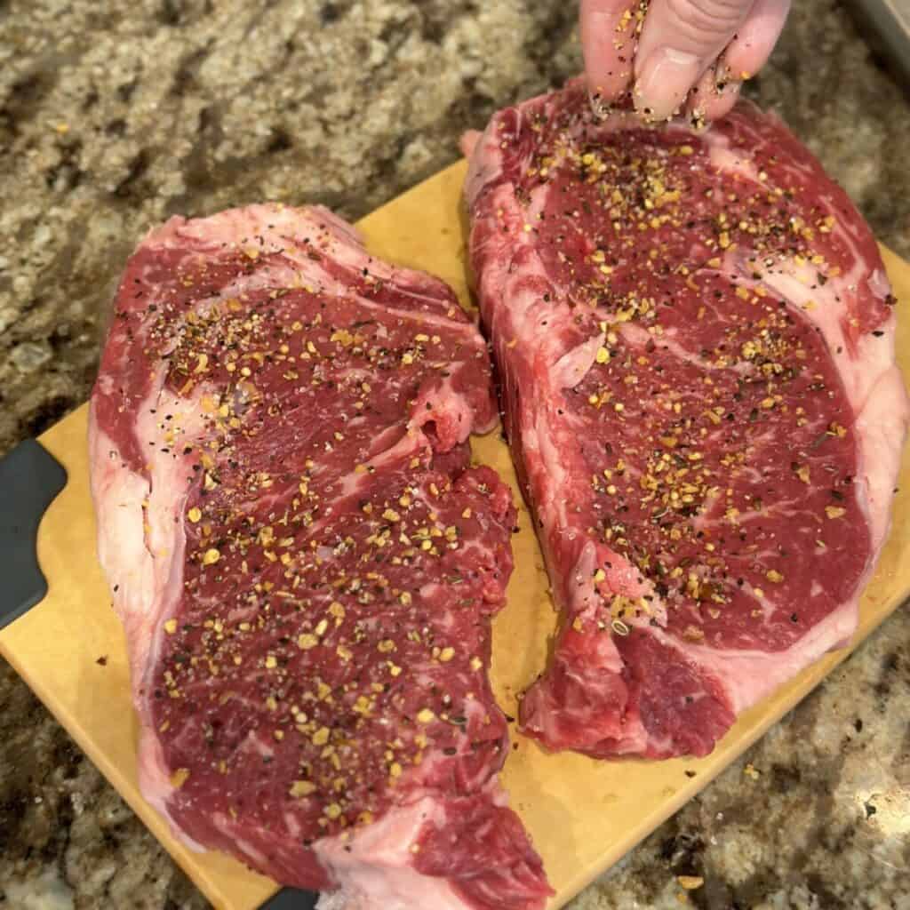 Seasoning ribeyes on a cutting board.