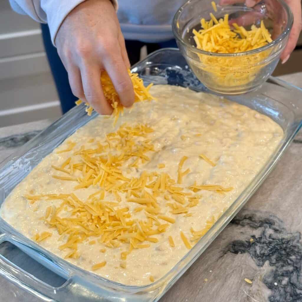 Adding cheese to the top of cheesy corn casserole in a baking dish.